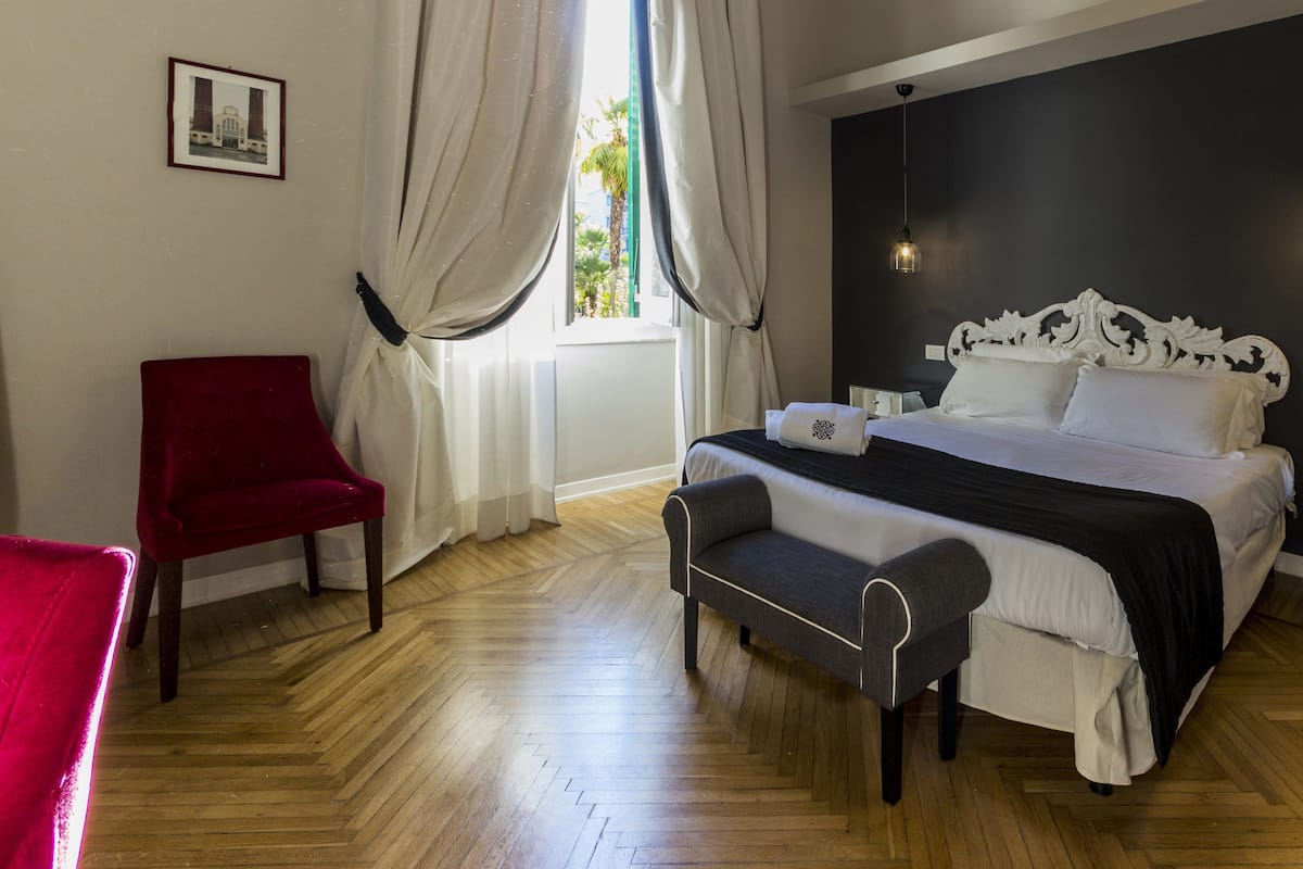 A bedroom with a bed, a red chair, a bench, a window with curtains, and a wall-mounted picture.