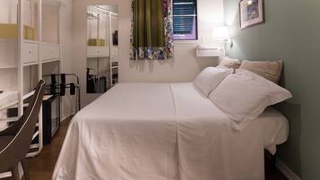 A bedroom with a bed, a wardrobe, a window with curtains, a bedside lamp, and a framed picture on the wall.