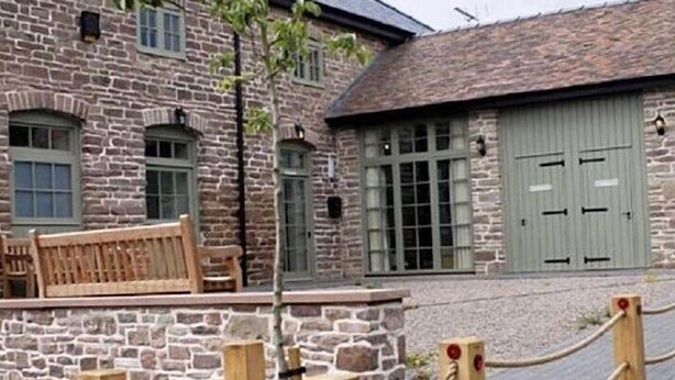 A stone building with a wooden bench, a large door, and a rope barrier. - England's Gate and Coachhouse (Hereford)