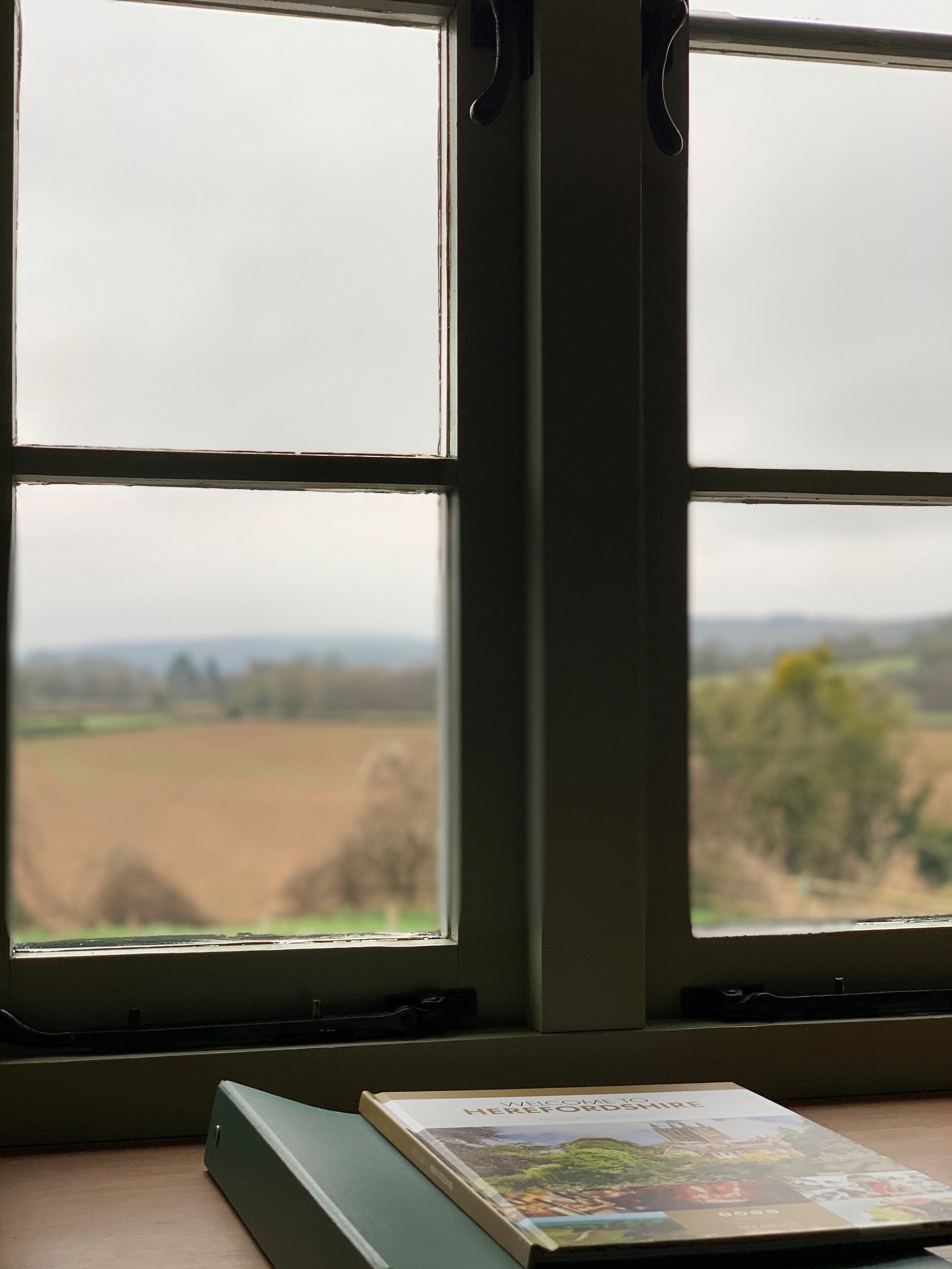 Et vindue med udsigt over et landligt landskab, en bog med titlen "Hereford" og et træbord.