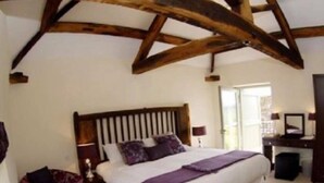 A bedroom with a wooden ceiling, a large bed with a headboard, a nightstand, a chair, and a desk with a lamp. - England's Gate and Coachhouse (Hereford)