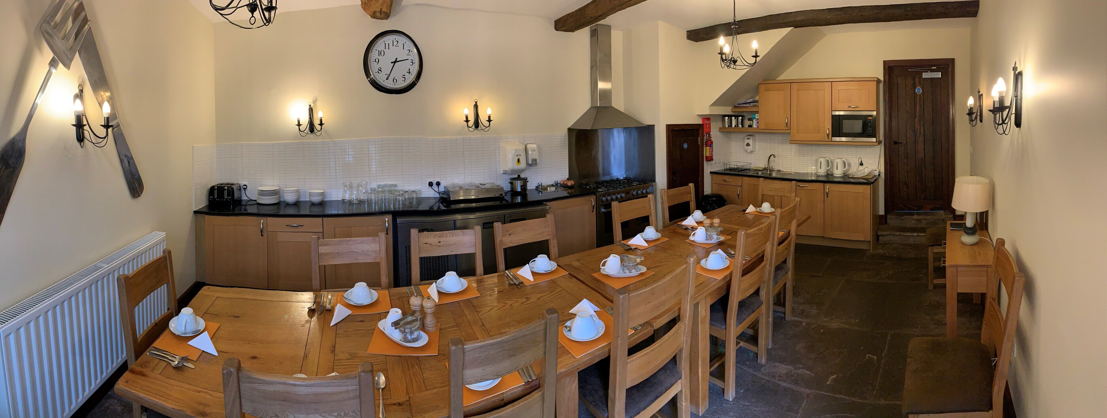 A kitchen with a stove, microwave, and wooden cabinets. There is a dining table set with white dishes and a clock on the wall.
