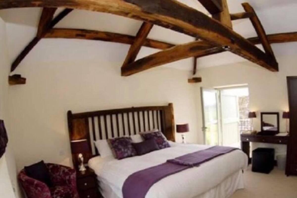 A bedroom with a wooden ceiling, a bed with white bedding and purple accents, a nightstand, and a chair.