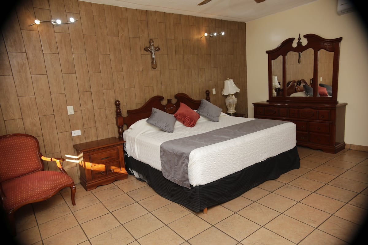 Un dormitorio con una cama de cabecera de madera, una mesita de noche de madera, una silla, un aparador y un espejo.