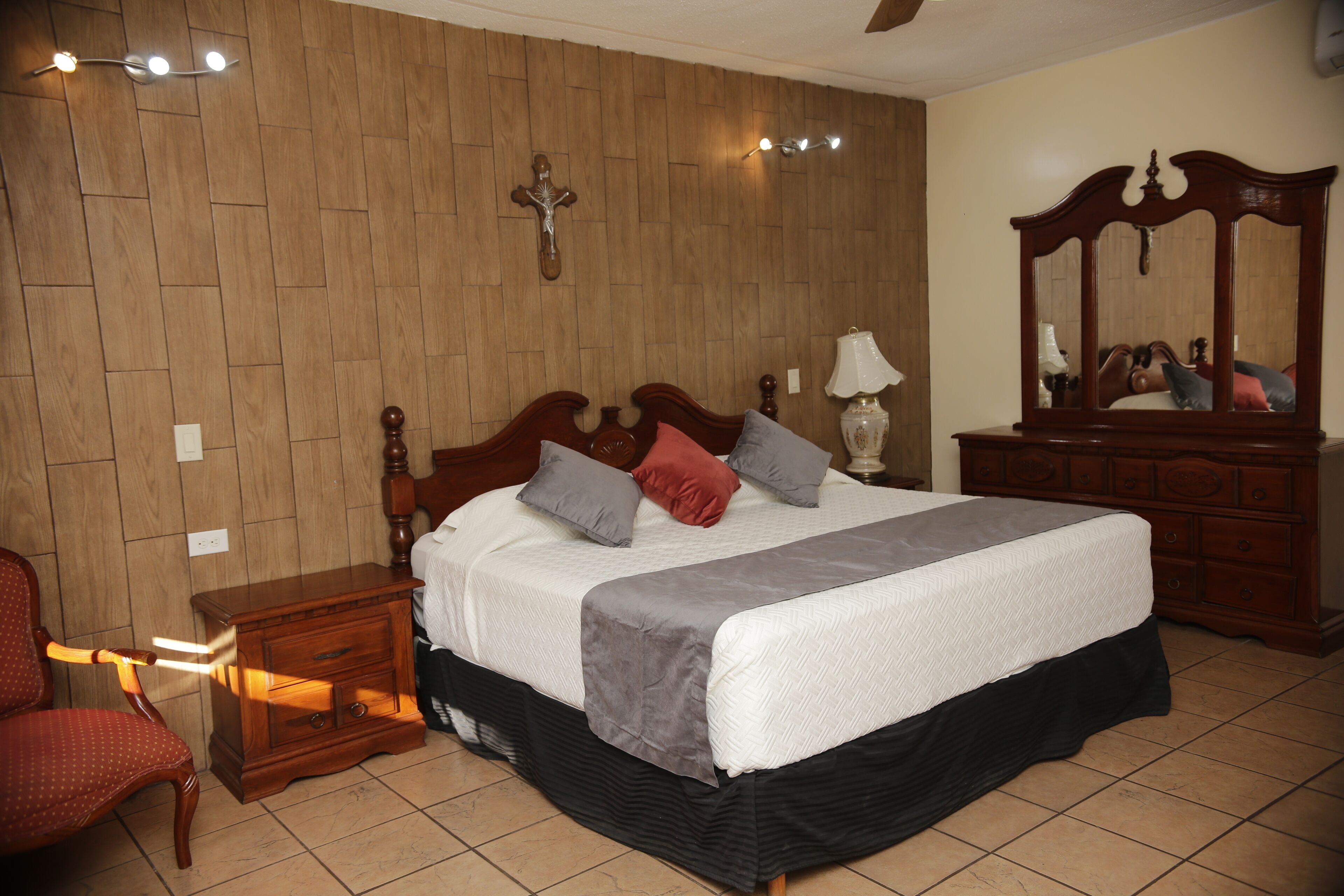 Une chambre à coucher avec une tête de lit en bois, un lit recouvert d’une couvre-lit blanche et grise, une table de chevet en bois, une chaise rouge et une commode en bois avec un miroir.