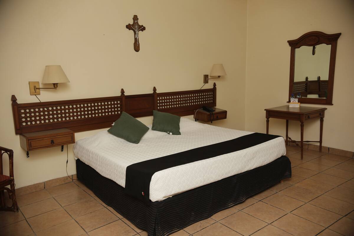 Un dormitorio con una cama de madera, dos mesitas de noche, un tocador, un espejo y una cruz colgada en la pared.