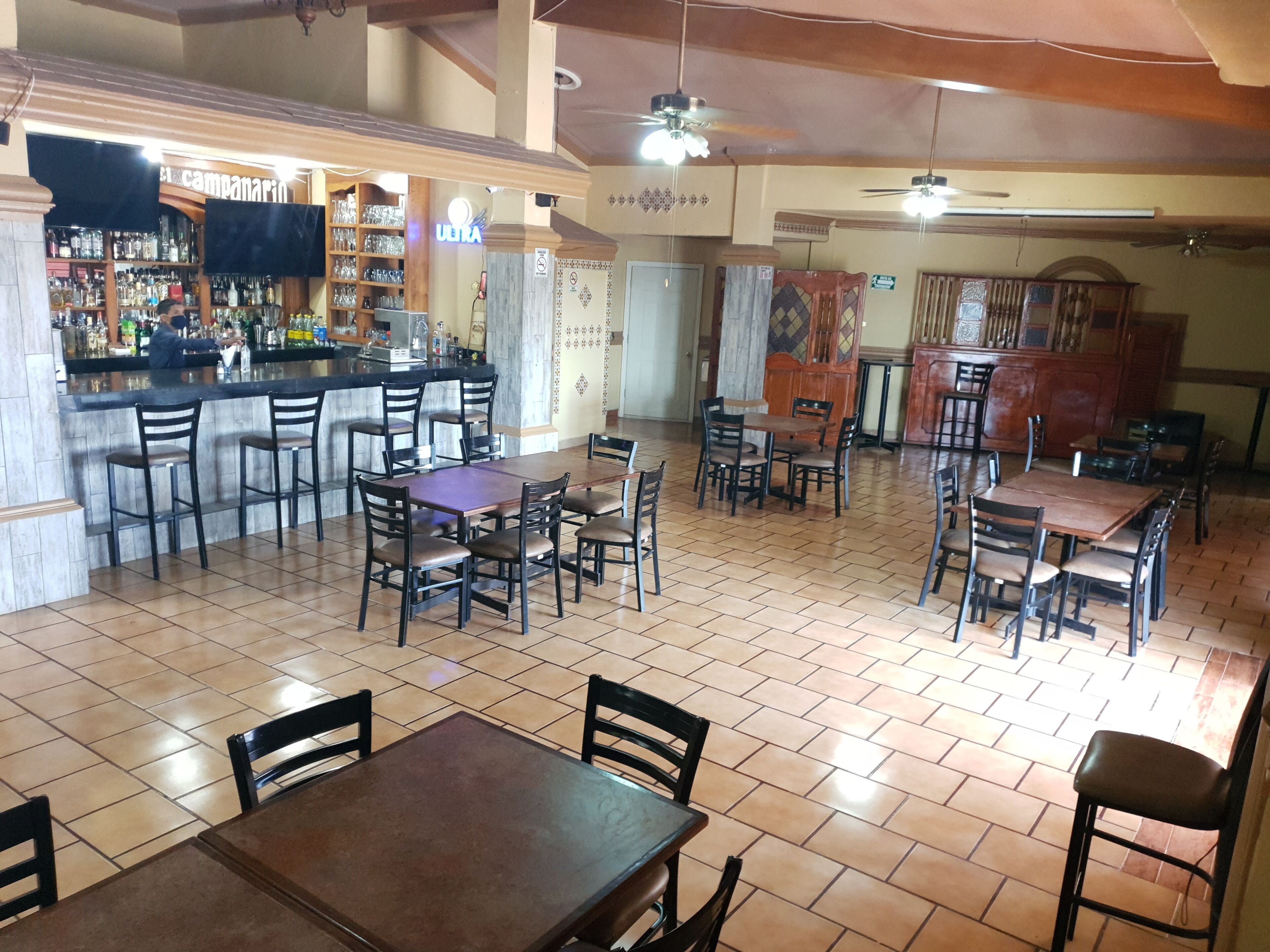 A restaurant with multiple dining tables and chairs, a bar area with shelves of bottles, and ceiling fans.