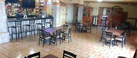 A restaurant with multiple dining tables and chairs, a bar area with shelves of bottles, and ceiling fans.