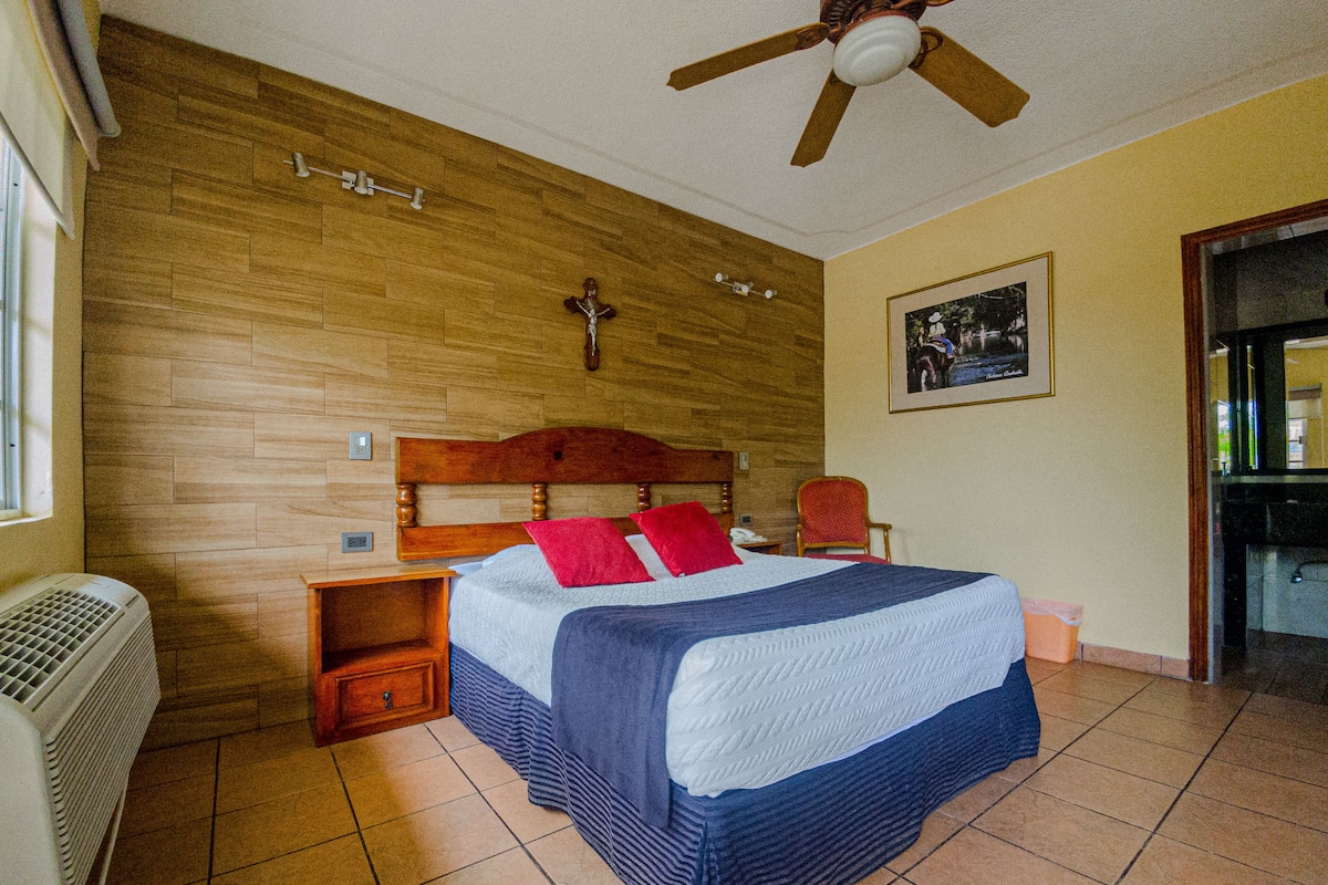 Habitación de hotel con cabecera de madera, cama con colcha azul y blanca, almohada roja, mesita de noche de madera, silla roja, ventilador de techo, cruz colgada en la pared y un cuadro enmarcado.