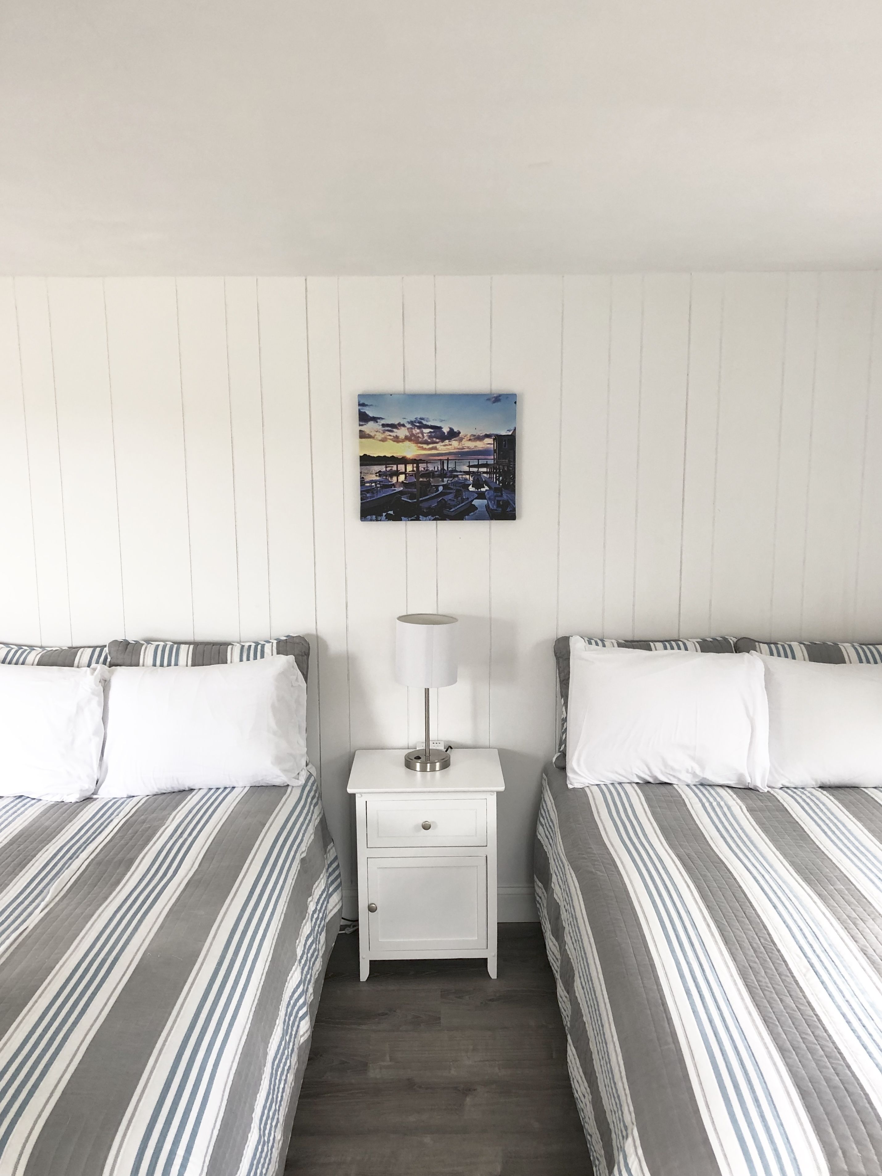 Una habitación de hotel con dos camas, una mesita de noche con lámpara y un cuadro con un paisaje al atardecer.