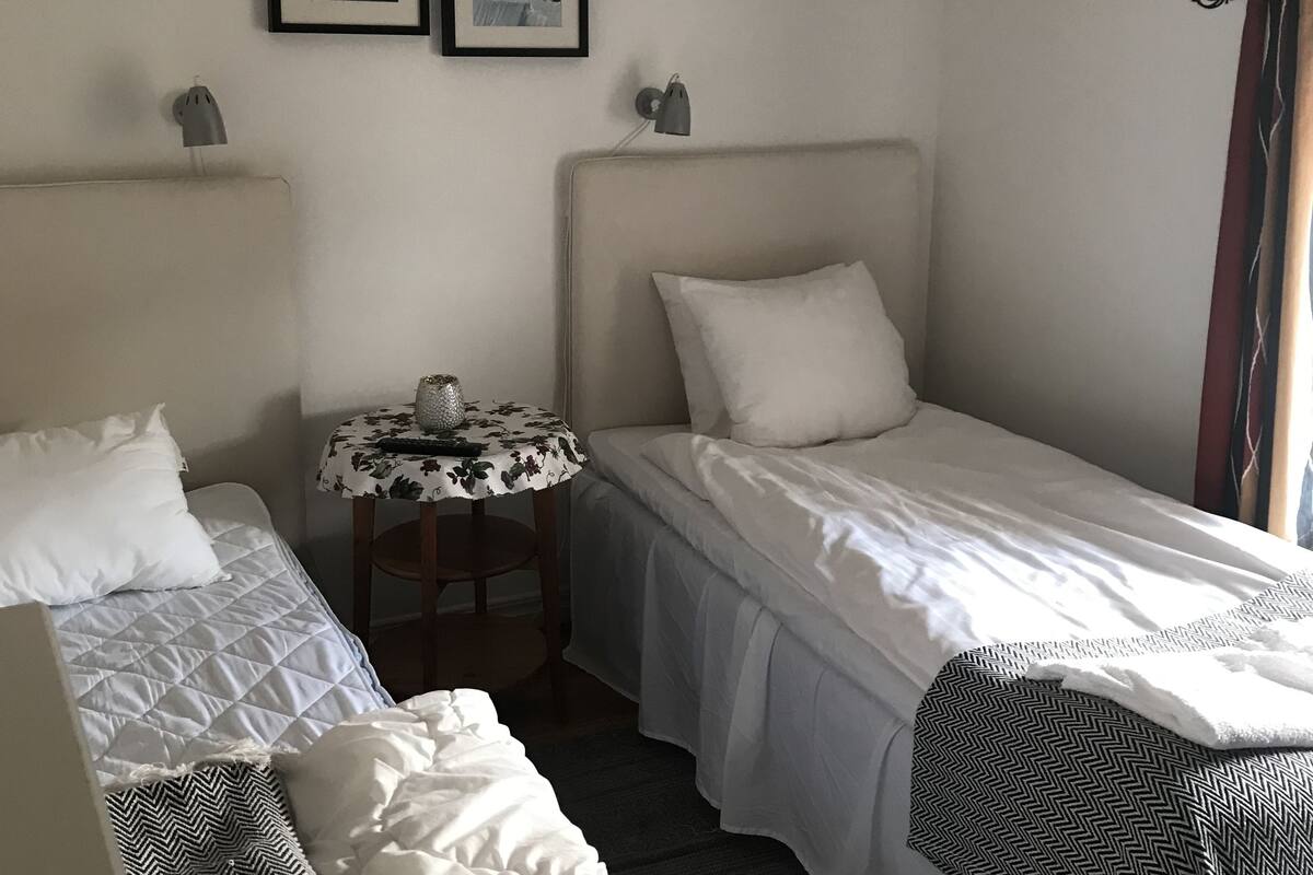 A bedroom with two single beds, a small table with a lamp, and two framed pictures on the wall.