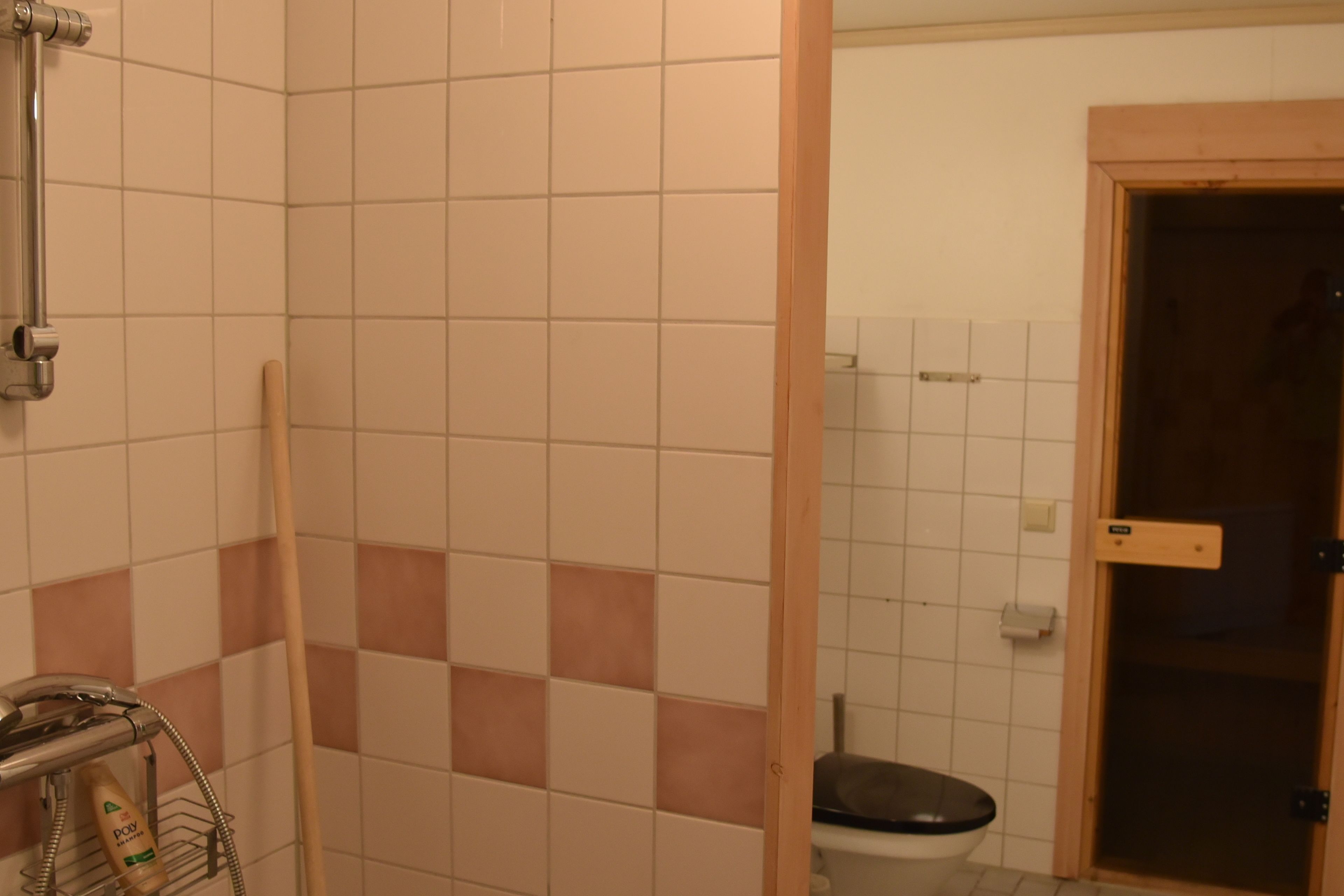 A bathroom with white and pink tiles, a shower, a toilet, and a wooden door.