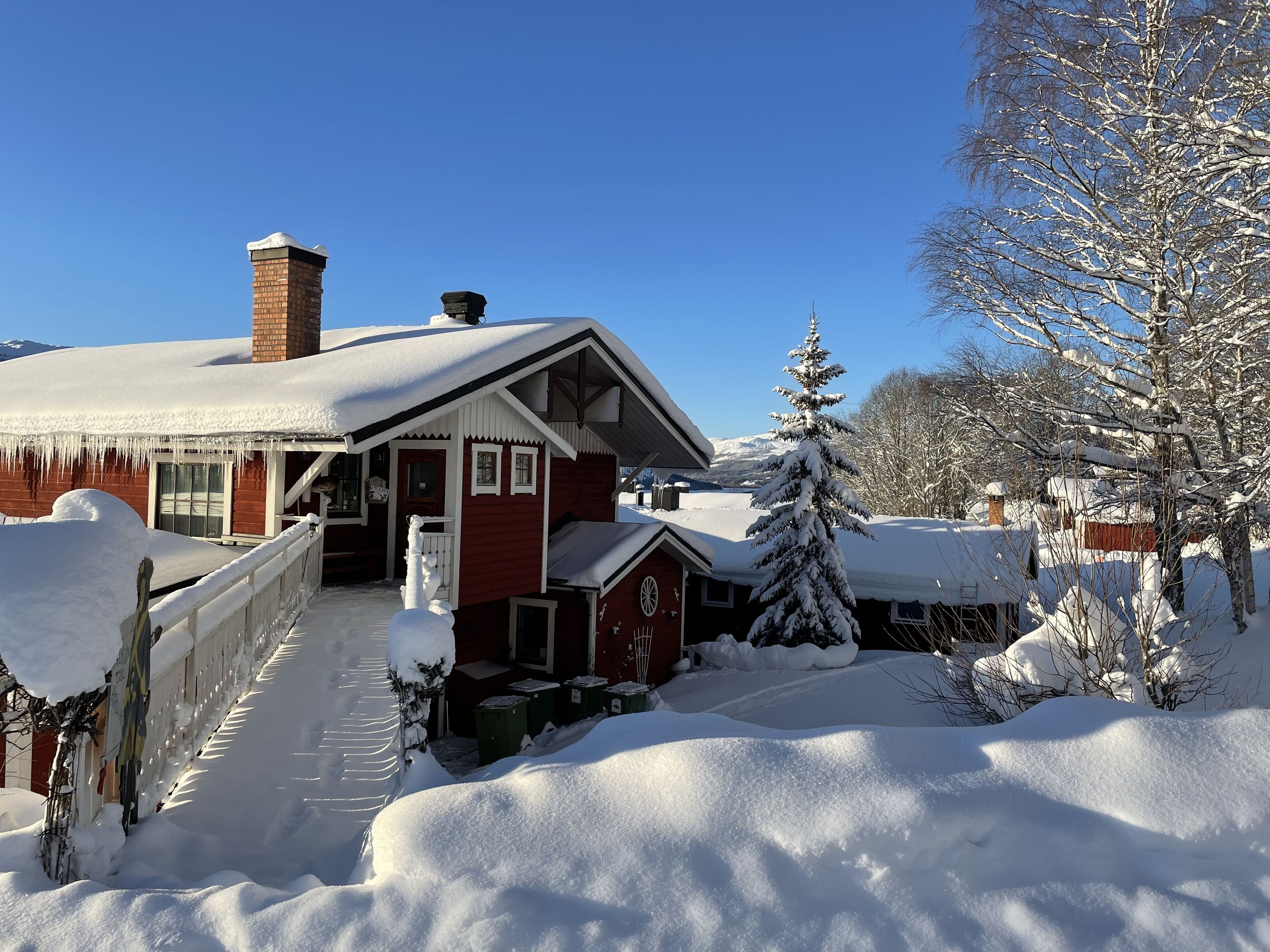 Ein mit Schnee bedecktes Holzhaus mit Schornstein, ein verschneiter Baum und ein klarer, blauer Himmel.