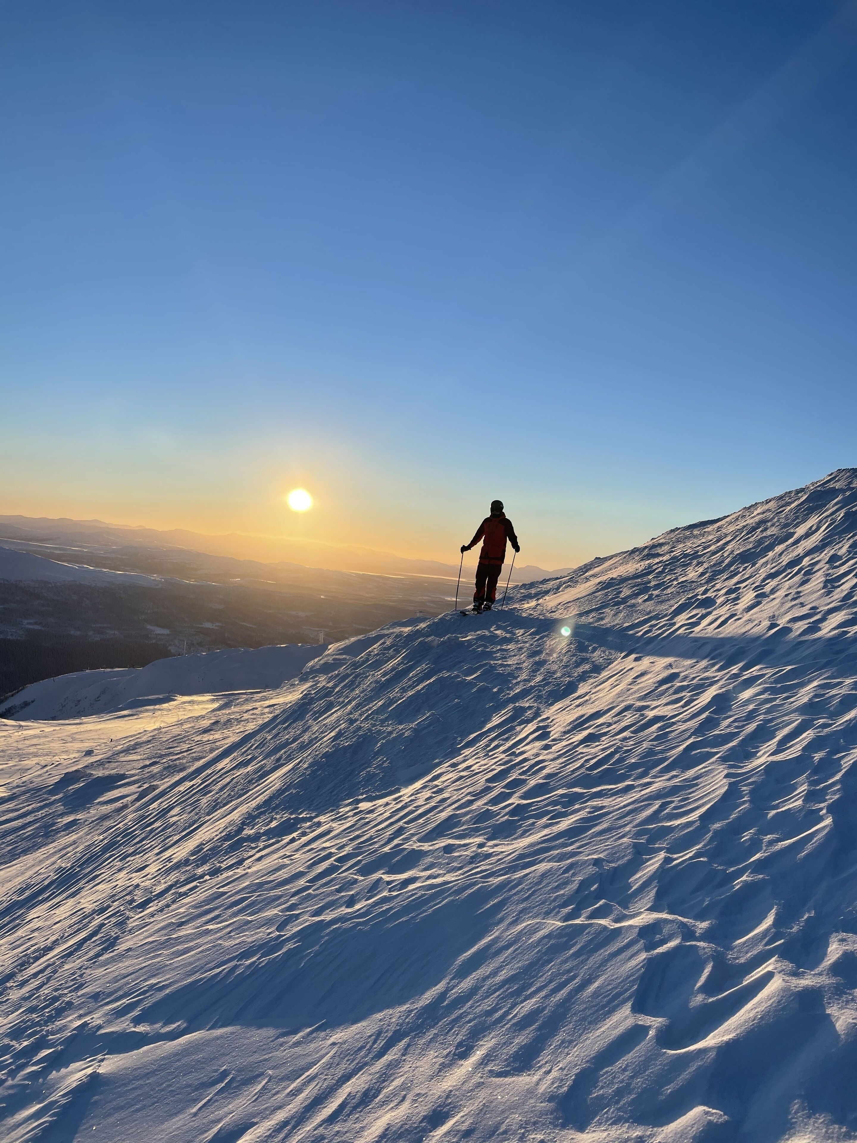 Una persona esquiando por una pendiente nevada bajo un cielo despejado, con el sol poniéndose en el horizonte.
