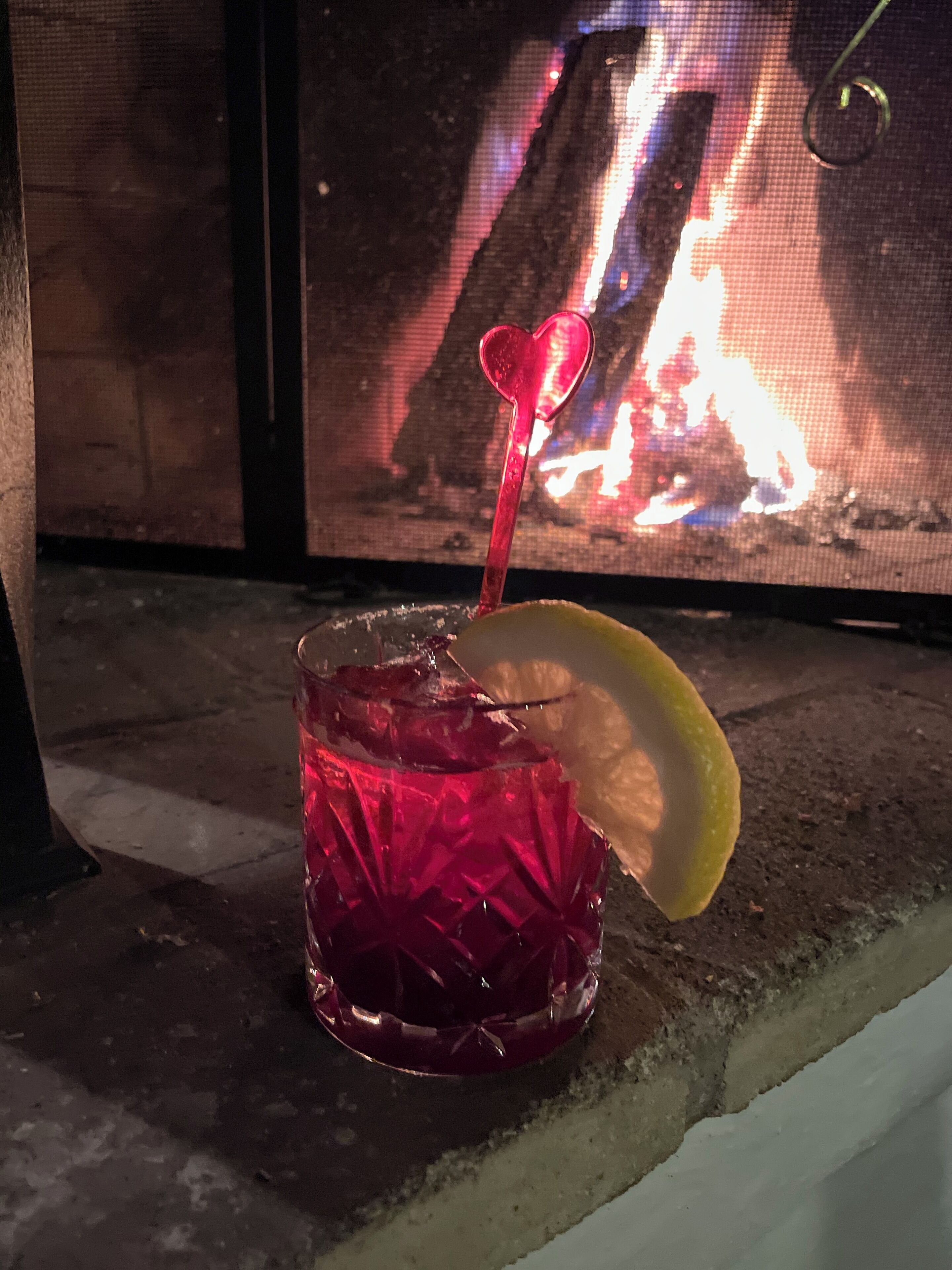 Un vaso de bebida roja con pajita en forma de corazón y una rodaja de limón, sobre una superficie texturizada con un espectáculo de fuego al fondo.