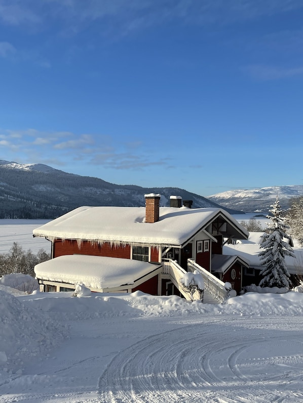 A snow-covered house with a chimney, a porch, and a view of a snowy landscape and mountains in the background.