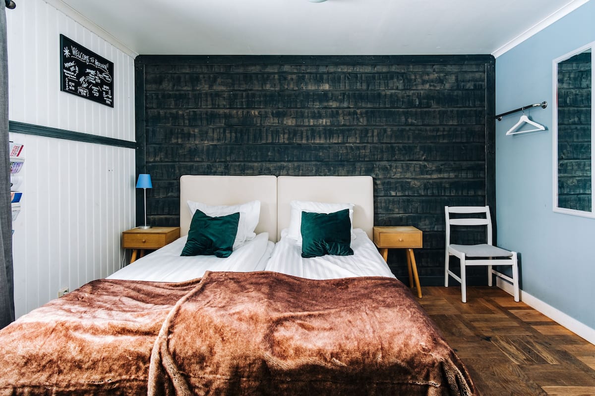 A bedroom with a brick-patterned wall, a bed with white bedding and green pillows, a brown blanket, a wooden nightstand with a lamp, and a white chair.