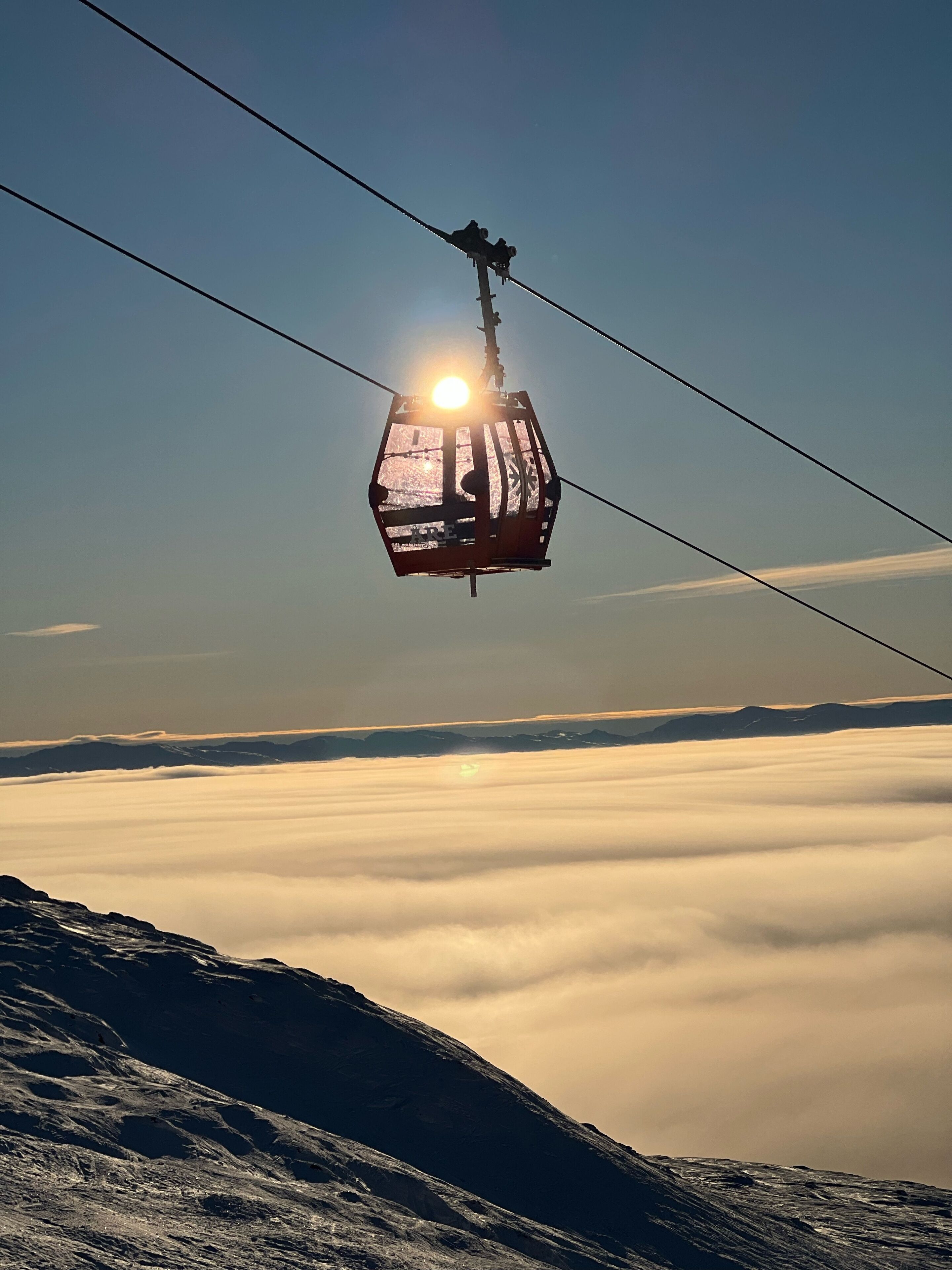 A cable car suspended above a mountain range with a clear sky and clouds below.