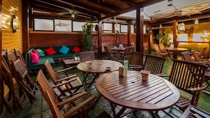 Een buitenruimte met houten tafels en stoelen, een groene bank met kleurrijke kussens en een houten plafondconstructie.