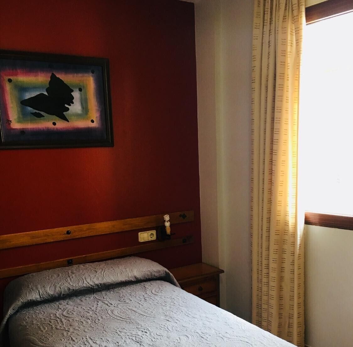 A hotel room with a bed, a nightstand, a wall clock, and a window with curtains.