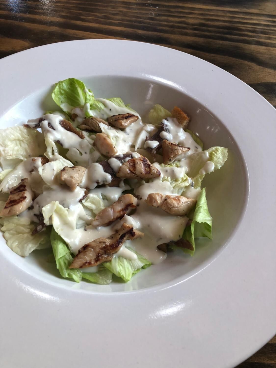 Een wit bord met een salade van sla, gegrilde kip en croutons, overgoten met een romige dressing.