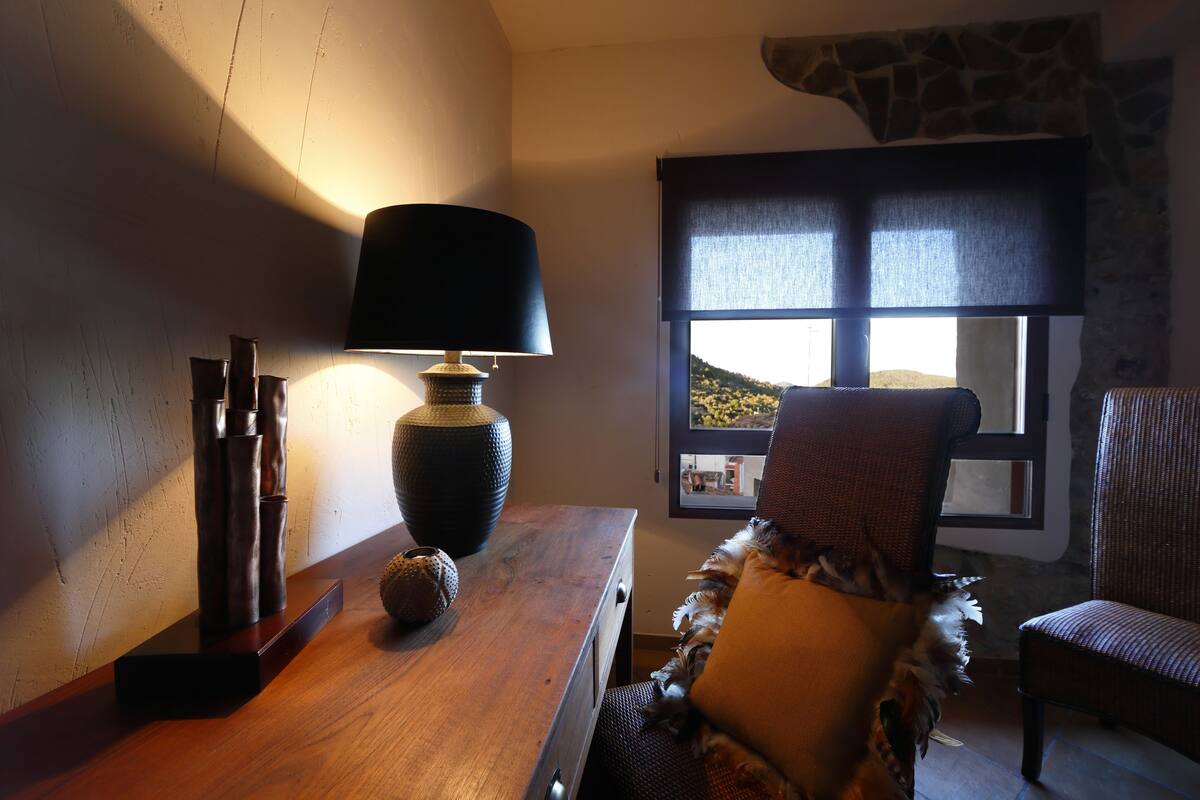 A room with a wooden desk, a lamp, a chair, and a window with a view of a hill.