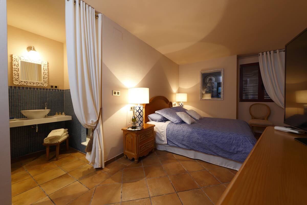A bedroom with a bed, a nightstand with a lamp, a dresser, a bathroom with a sink and mirror, and a window with curtains.