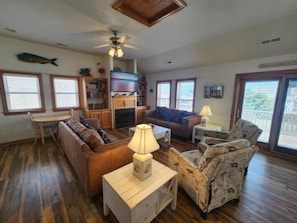 A living room with a sofa, armchair, wooden coffee table, and a TV. There are bookshelves, a ceiling fan, and a large window. - Free Private Heated Pool, Hot Tub, Fall flex stays, Pets OK, Rec. Room, Close to Beach & Restaura... (Kill Devil Hills)