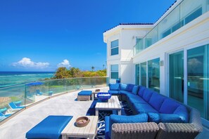 A modern outdoor living area with a large blue sofa, wicker ottomans, and a glass balustrade overlooking the ocean.