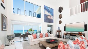 Amplia sala de estar con un gran ventanal con vista al océano, un sofá blanco con cojines estampados, una mesa de centro de madera y un televisor de pantalla plana sobre un mueble de madera.