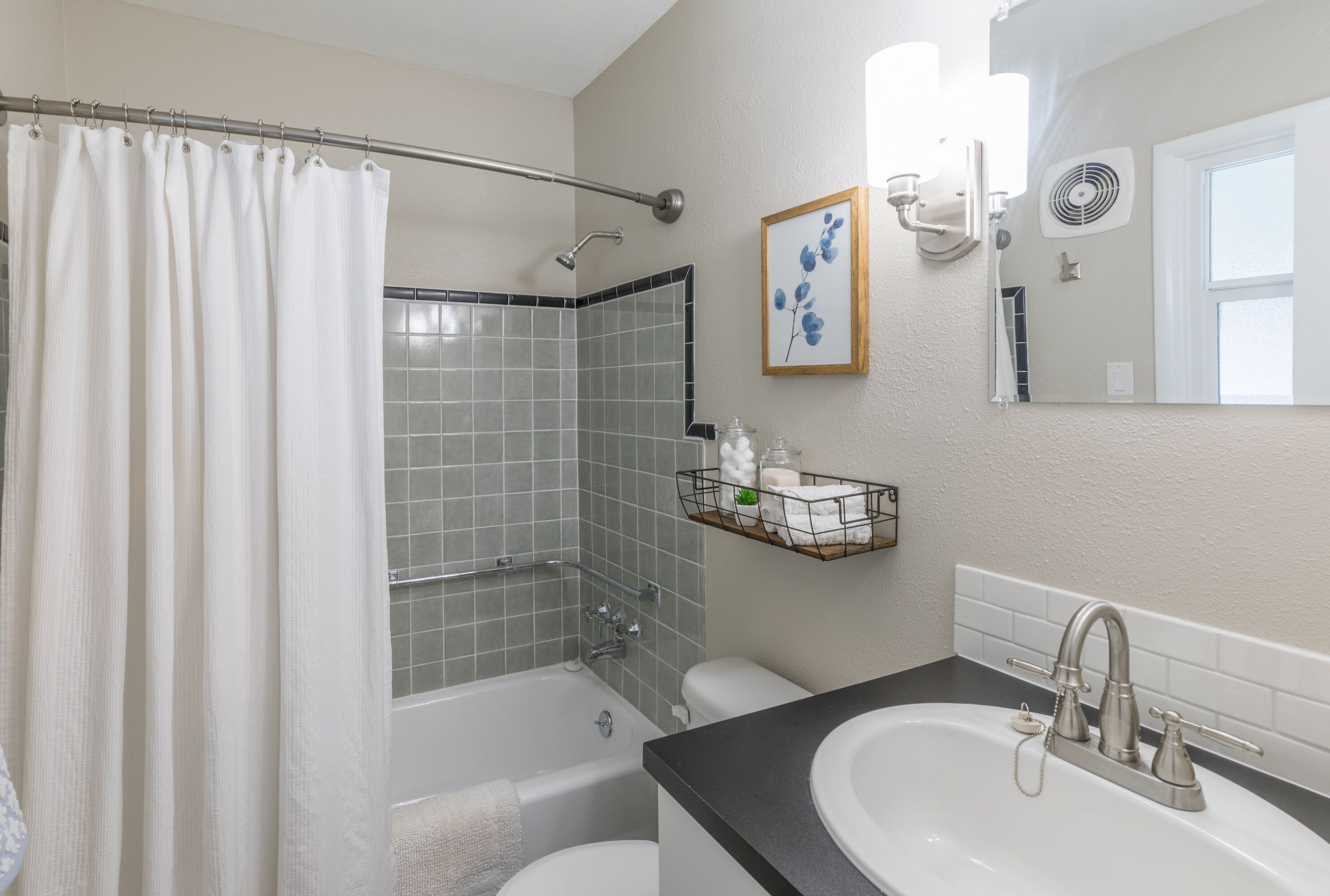 Une salle de bain avec une baignoire, des toilettes et un lavabo. On y trouve un rideau de douche, un porte-serviettes et une étagère avec des articles de toilette.