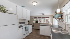 A modern kitchen with white cabinets, a stove, and a sink. There is a ceiling fan and a window with curtains.
