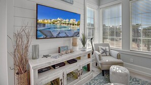 A living room with a large TV mounted on the wall, a comfortable seating area with a chair and ottoman, and a view of a pool and houses through the windows.