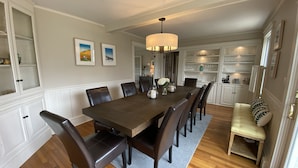 Une salle à manger avec une grande table en bois, des fauteuils en cuir foncé, un lustre et un canapé agrémenté de coussins à motifs.