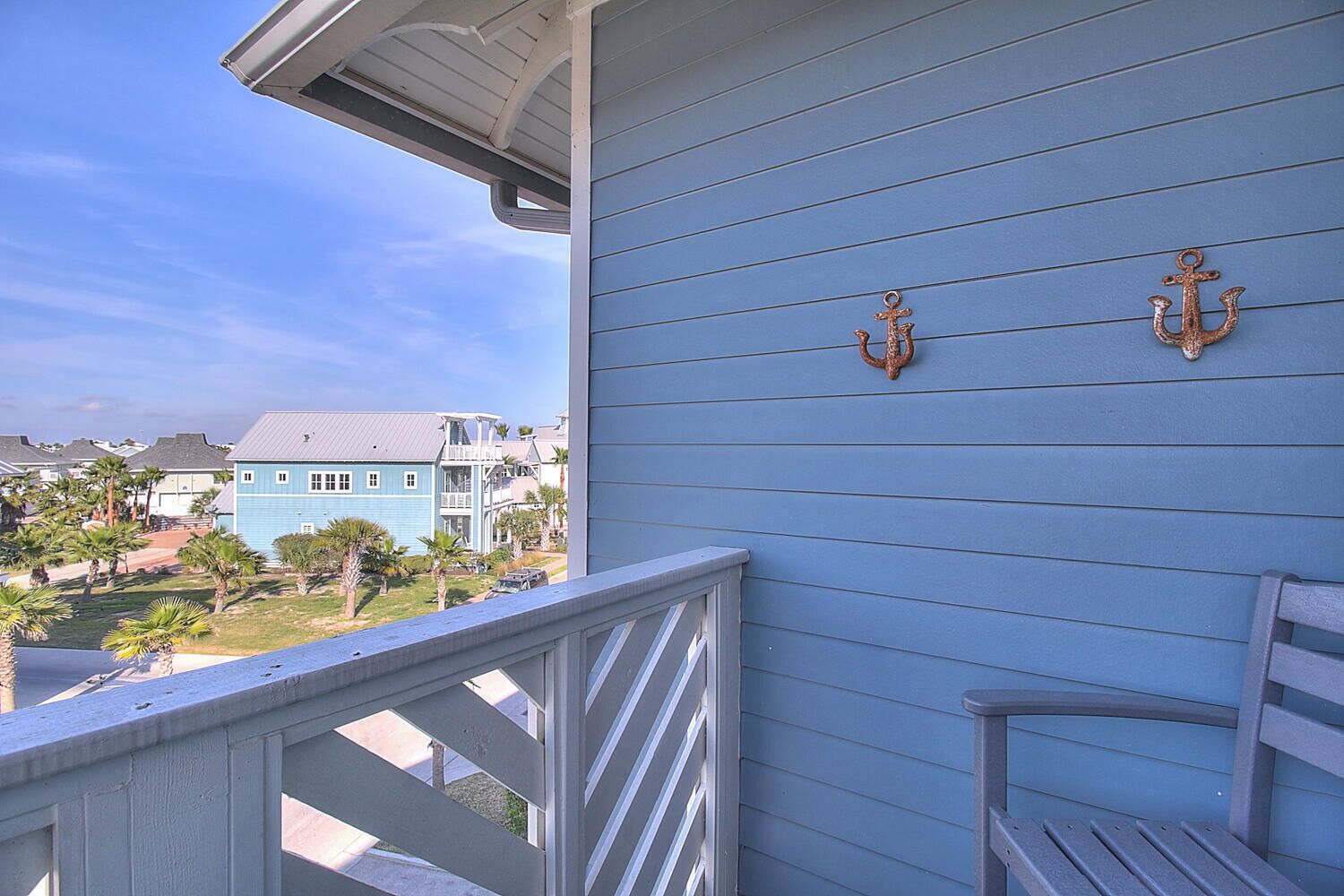 A balcony with a railing, a blue building with two anchor decorations, and a view of other houses and palm trees.