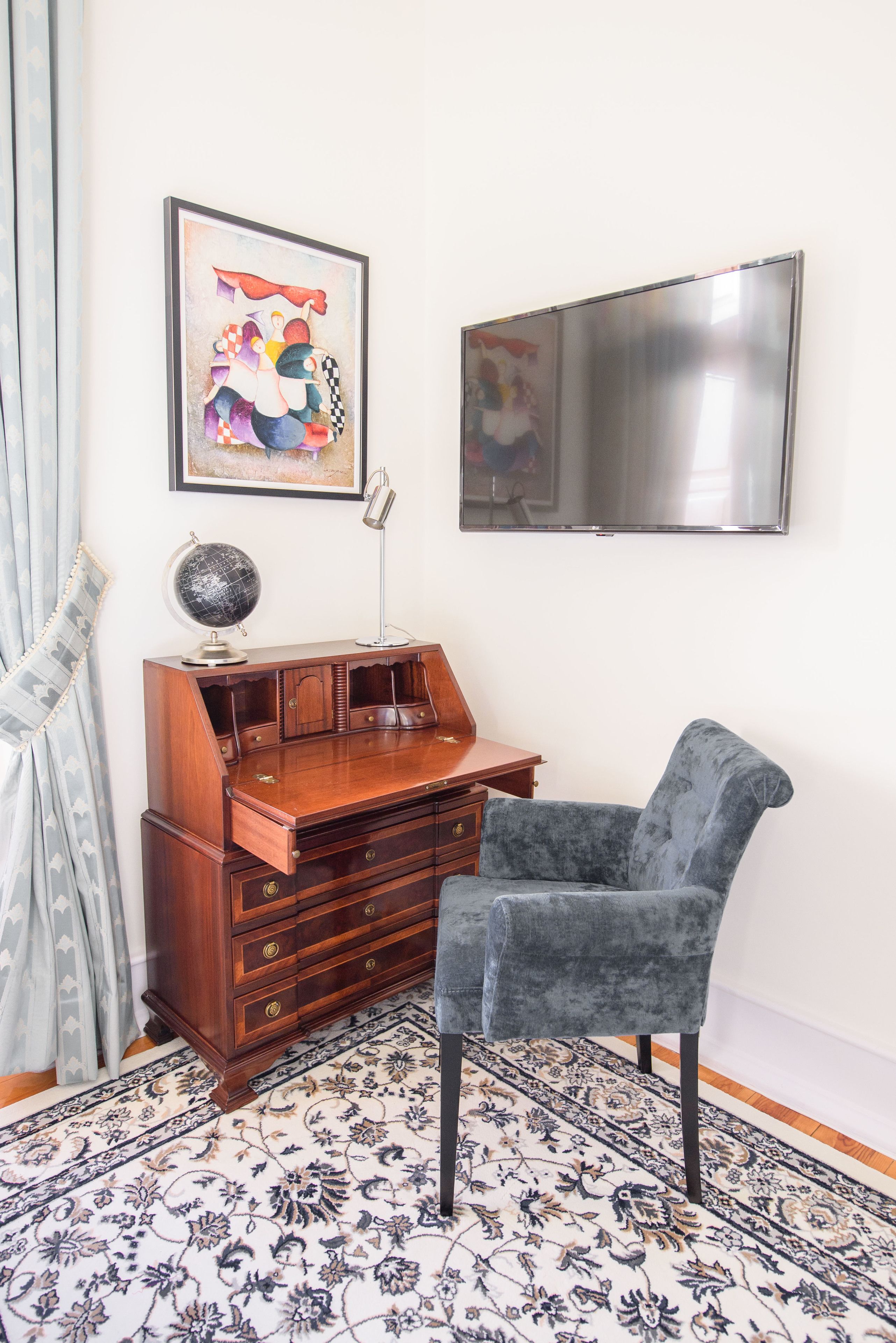Een kamer met een houten bureau, een wereldbol, een lamp, een grijze stoel, een kamerbreed tapijt met dessin en een schilderij aan de muur.