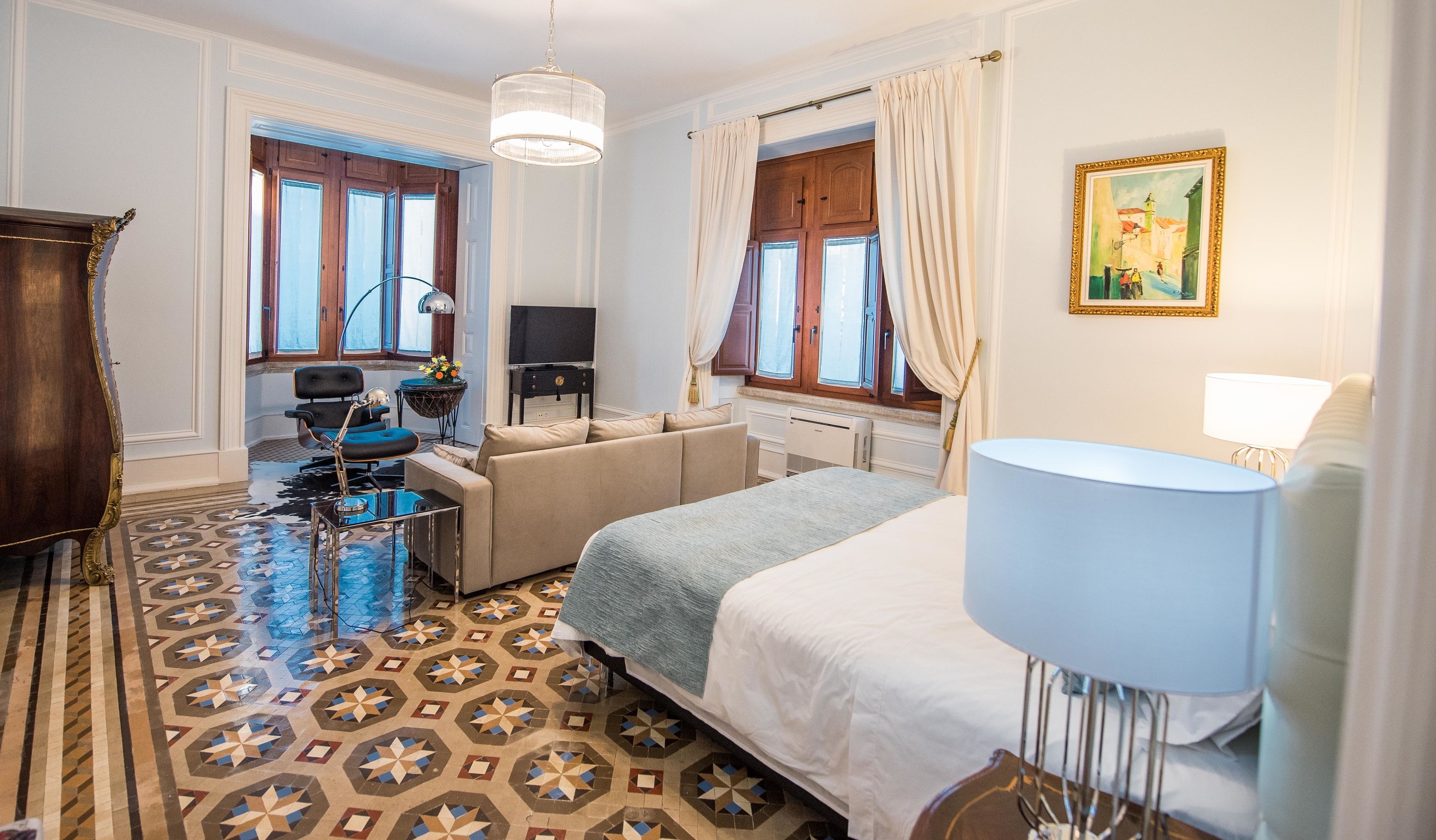 Habitación de hotel con cama, sofá, escritorio, silla, lámpara, televisor y ventana con cortinas.