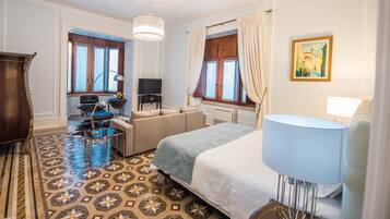 Habitación de hotel con cama, sofá, escritorio, silla, lámpara, televisor y ventana con cortinas.