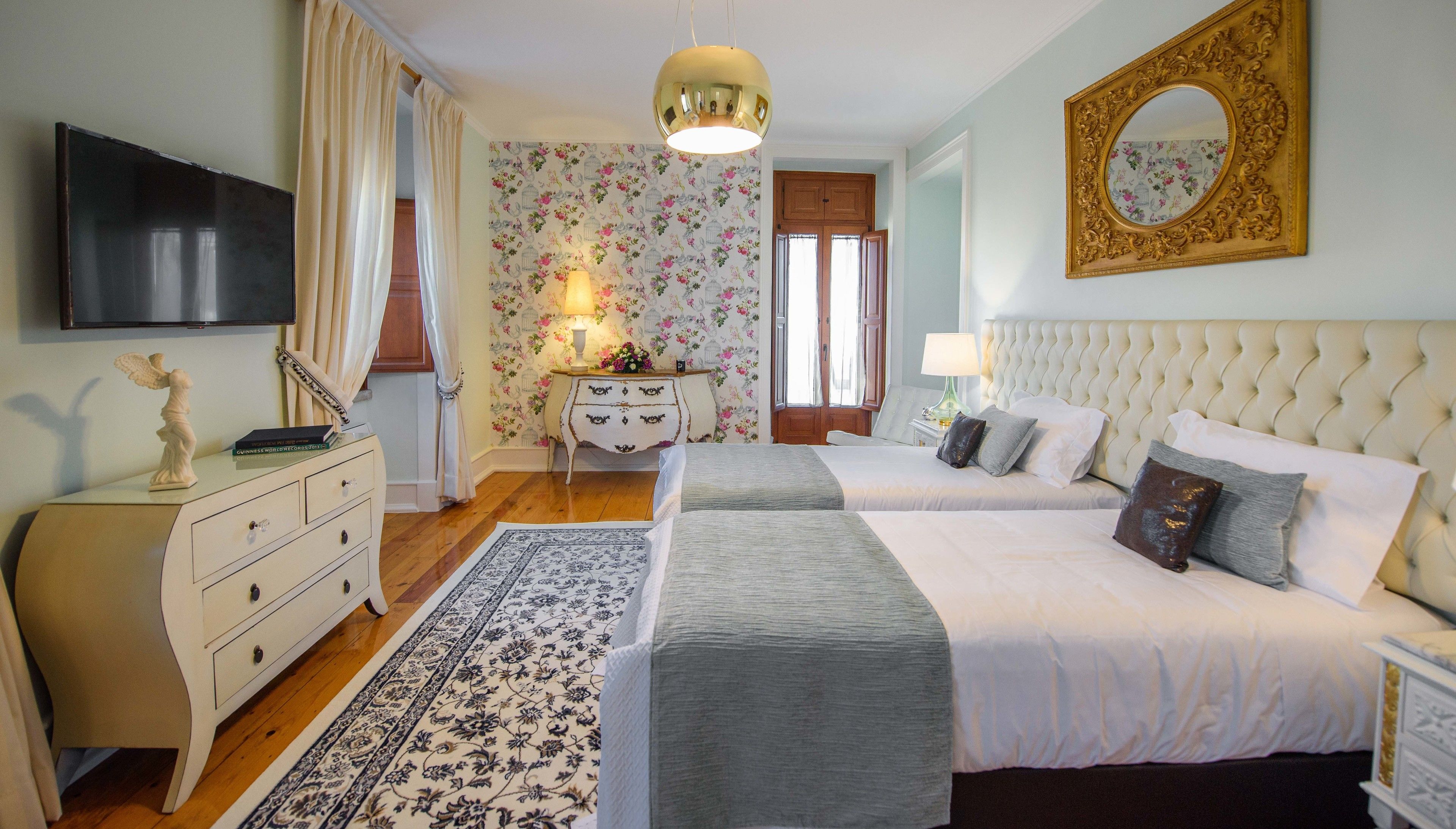A hotel room with two beds, a dresser, a television, a mirror, and a floral wallpaper.