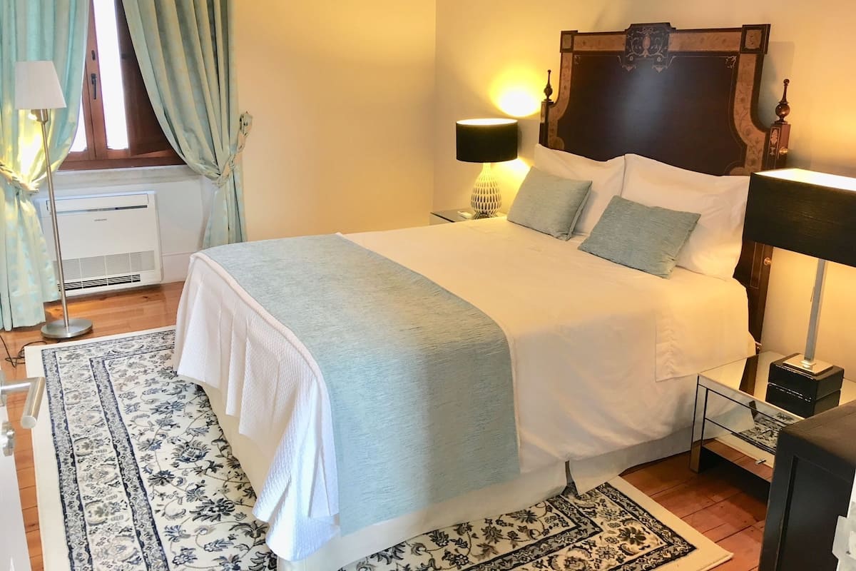 Un lit bien fait, recouvert d’une couvre-lit blanche et bleu clair, avec deux lampes de chevet, une petite table de chevet avec une lampe, une tête de lit décorative et un tapis à motifs sur le sol.