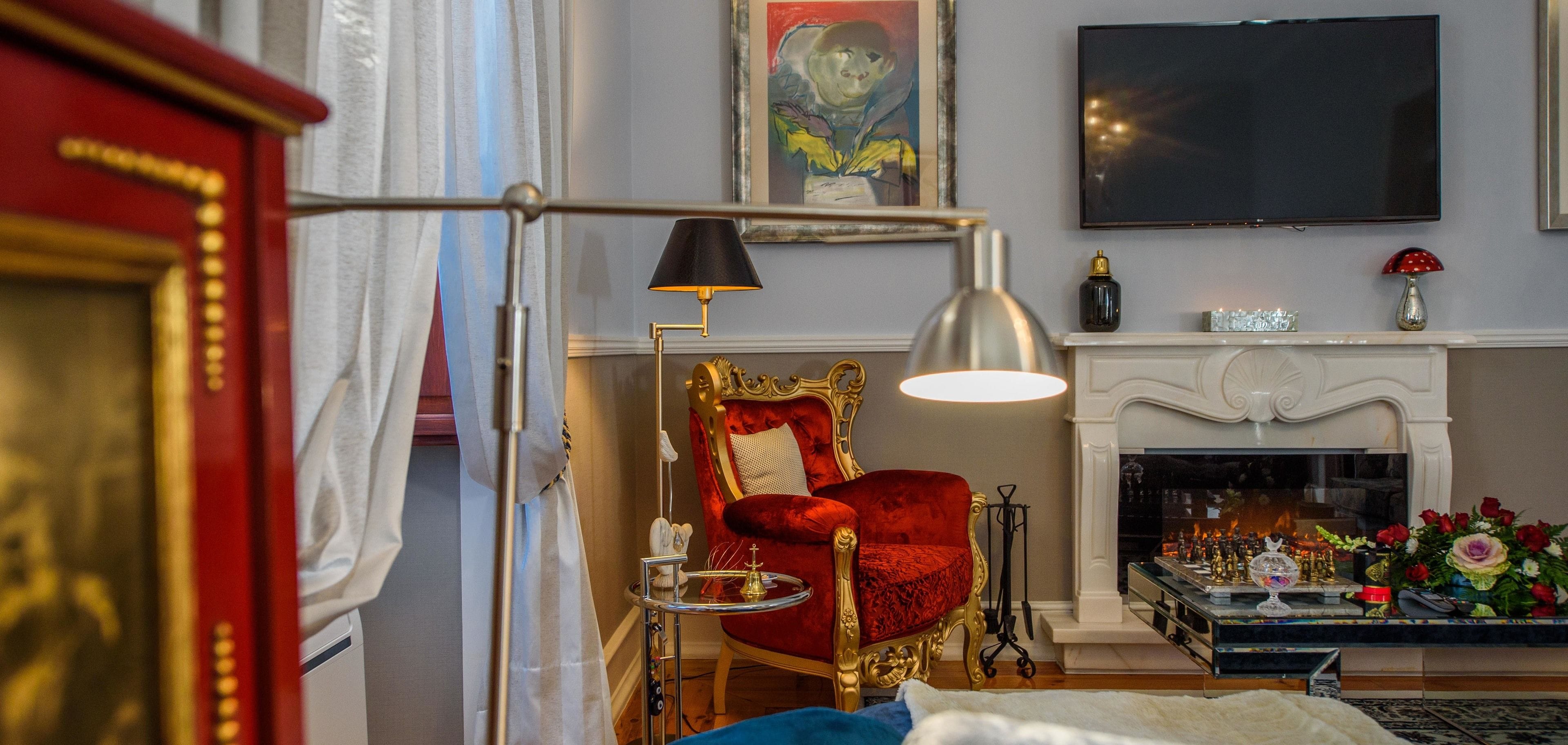 Une pièce avec un fauteuil en velours rouge, une cheminée blanche, un téléviseur à écran plat et un lustre.