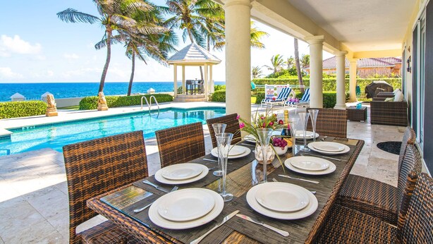 Un área de comedor junto a la piscina con mobiliario de mimbre, un pabellón y vista al océano.