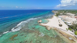 Vista aérea de una playa con aguas turquesas cristalinas, una orilla arenosa y algunos edificios al fondo.