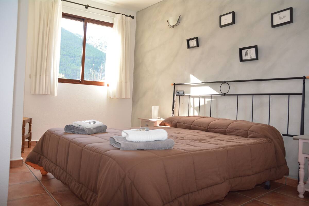 A bedroom with a large bed, a window with curtains, a bedside table with a lamp, and framed pictures on the wall.