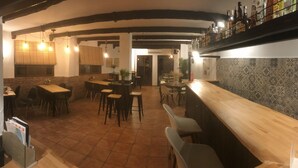 Zona de bar con mostrador y taburetes de madera, una cocina al fondo y suelo de baldosas.