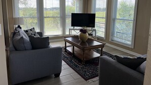 Un salon comprenant un canapé, une table basse avec un vase, un téléviseur à écran plat et de grandes fenêtres équipées de stores.