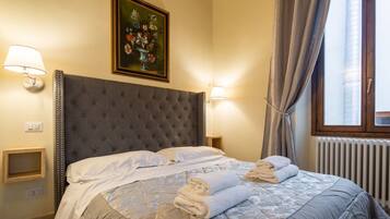 Ein Hotelzimmer mit einer geblümten Kopfteil, einem Gemälde über dem Bett, zwei Nachttischlampen, einem Fenster mit Vorhängen und einer Heizung.