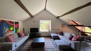 A loft-style living room with a high ceiling, exposed wooden beams, a large window with a view of greenery, a flat-screen TV, two sofas, a coffee table, and a colorful abstract painting on the wall. - Family Friendly Farmhouse -  Secluded 2 acres with Private Pool & Stream (Sassello)