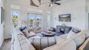 A spacious living room with a large sofa, a coffee table, and a fireplace. There are large windows and a ceiling fan.