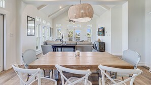 A spacious dining area with a wooden table and white wicker chairs, a large woven pendant light, and a view of the kitchen and living area through large windows.